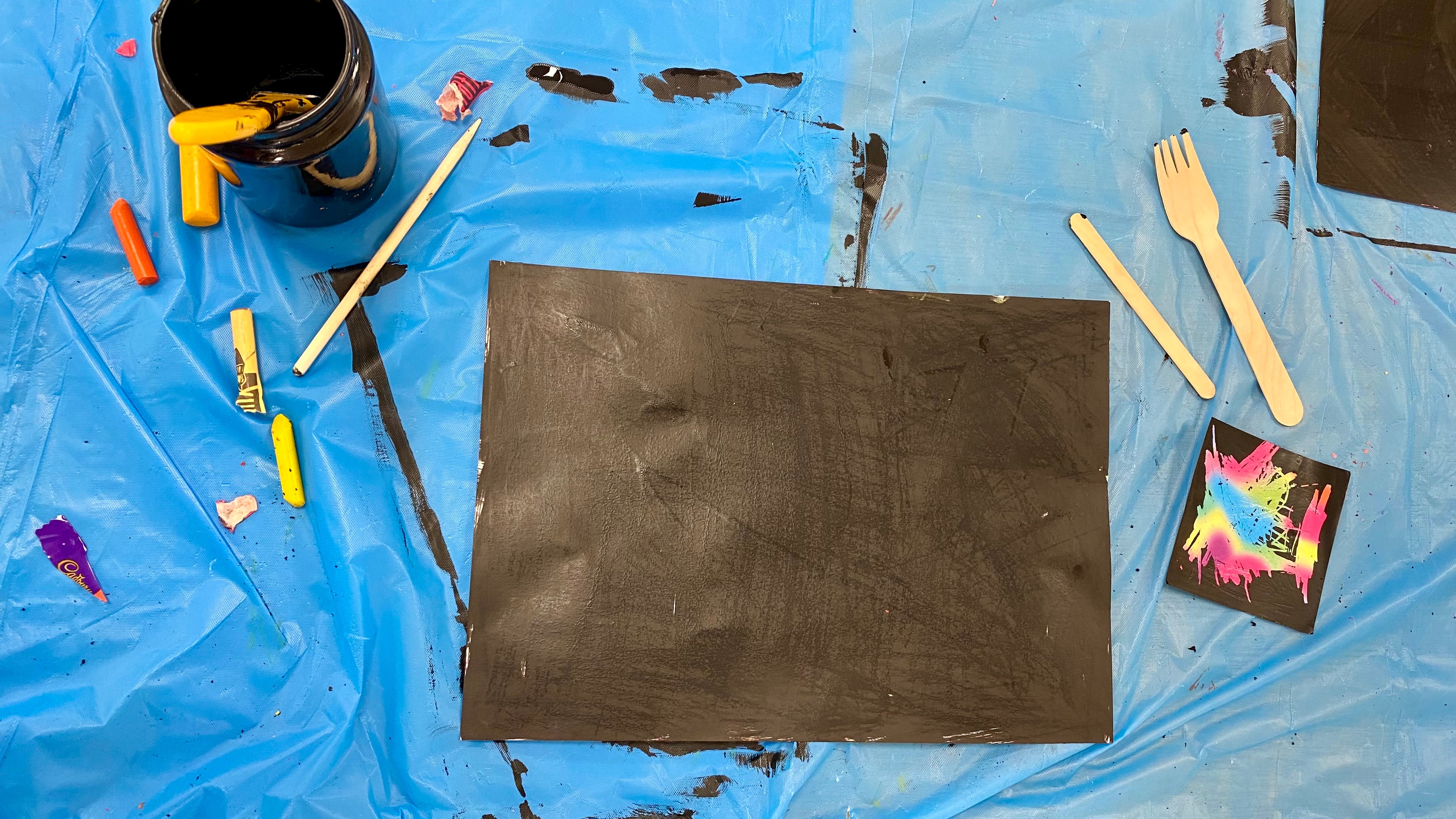 A sheet of black card on a tabletop surrounded by art supplies including black paint, scratch tools and crayons