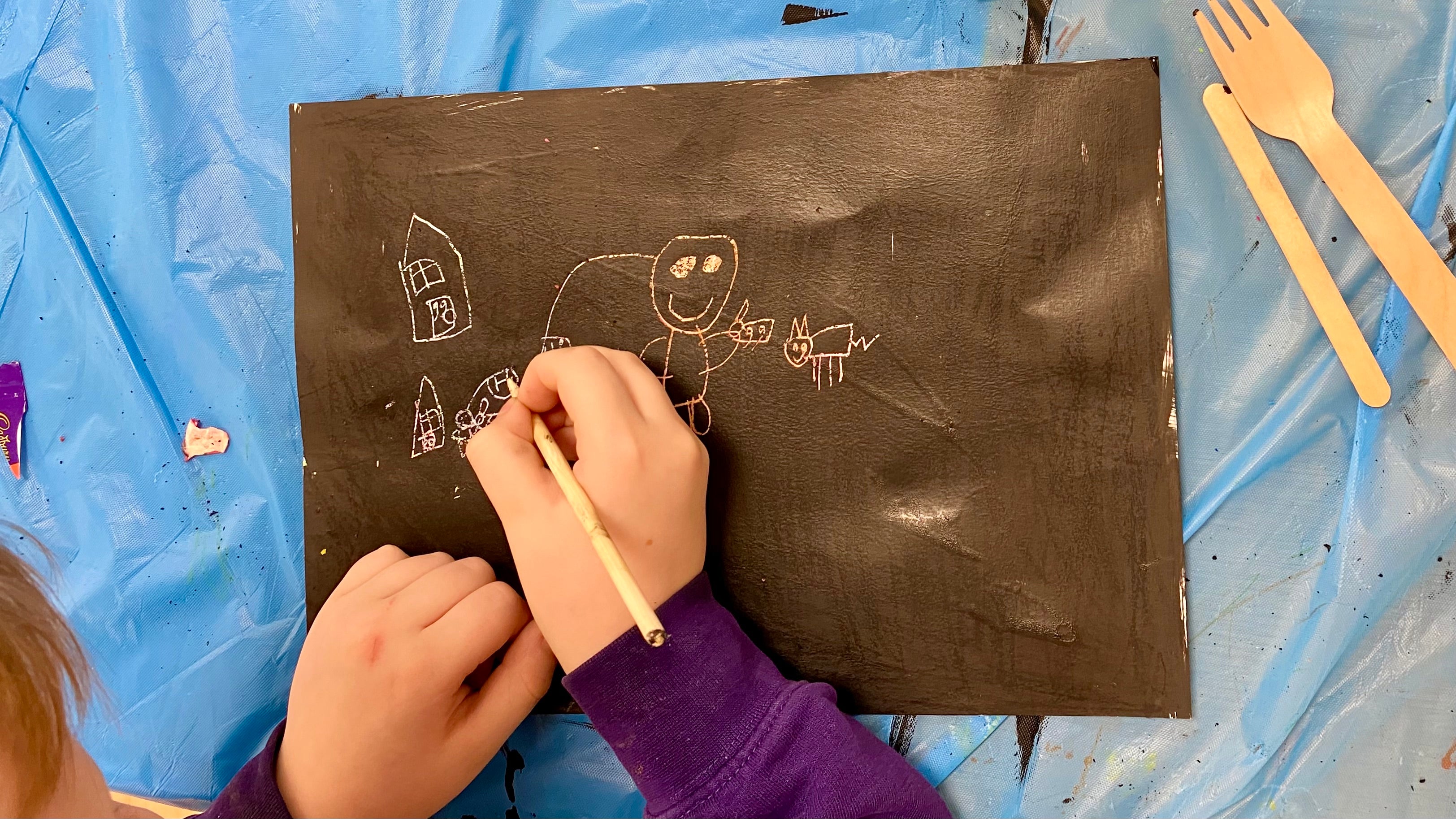 A hand holding a wooden scratching tool etching into a sheet of handmade scratch art paper. There is a line drawing of a person, a cat and some houses etched in to the black revealing bright colours beneath.