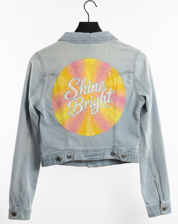Denim jacket with a colourful hand painted sunburst and 'Shine Bright' written in the centre.