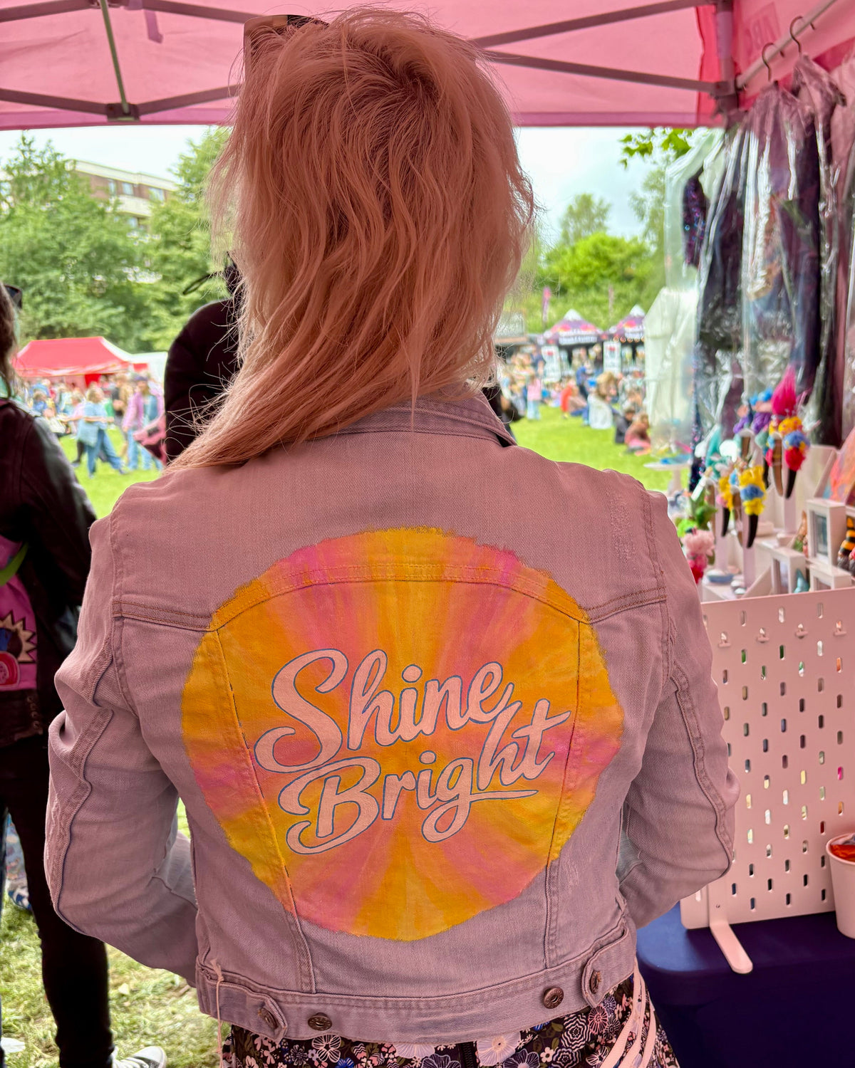 A person viewed from behind wearing a  pale denim jacket with a large circular painted back graphic that reads 'Shine Bright' in white script. The scene is at an outdoor festival under a pink canopy, with clothing stalls and grass visible in the background.