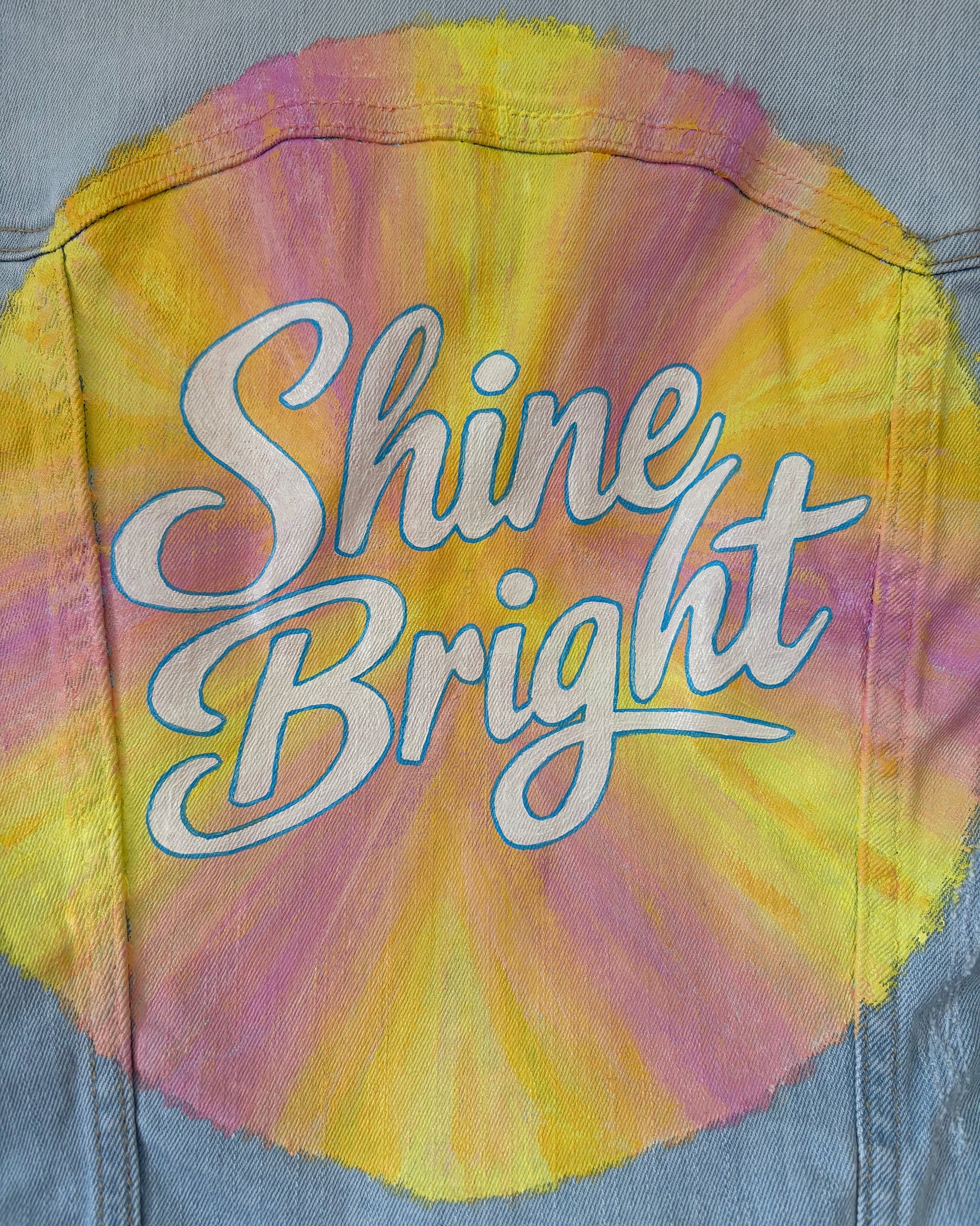 Close up of denim jacket with a colourful hand painted sunburst and 'Shine Bright' written in the centre.
