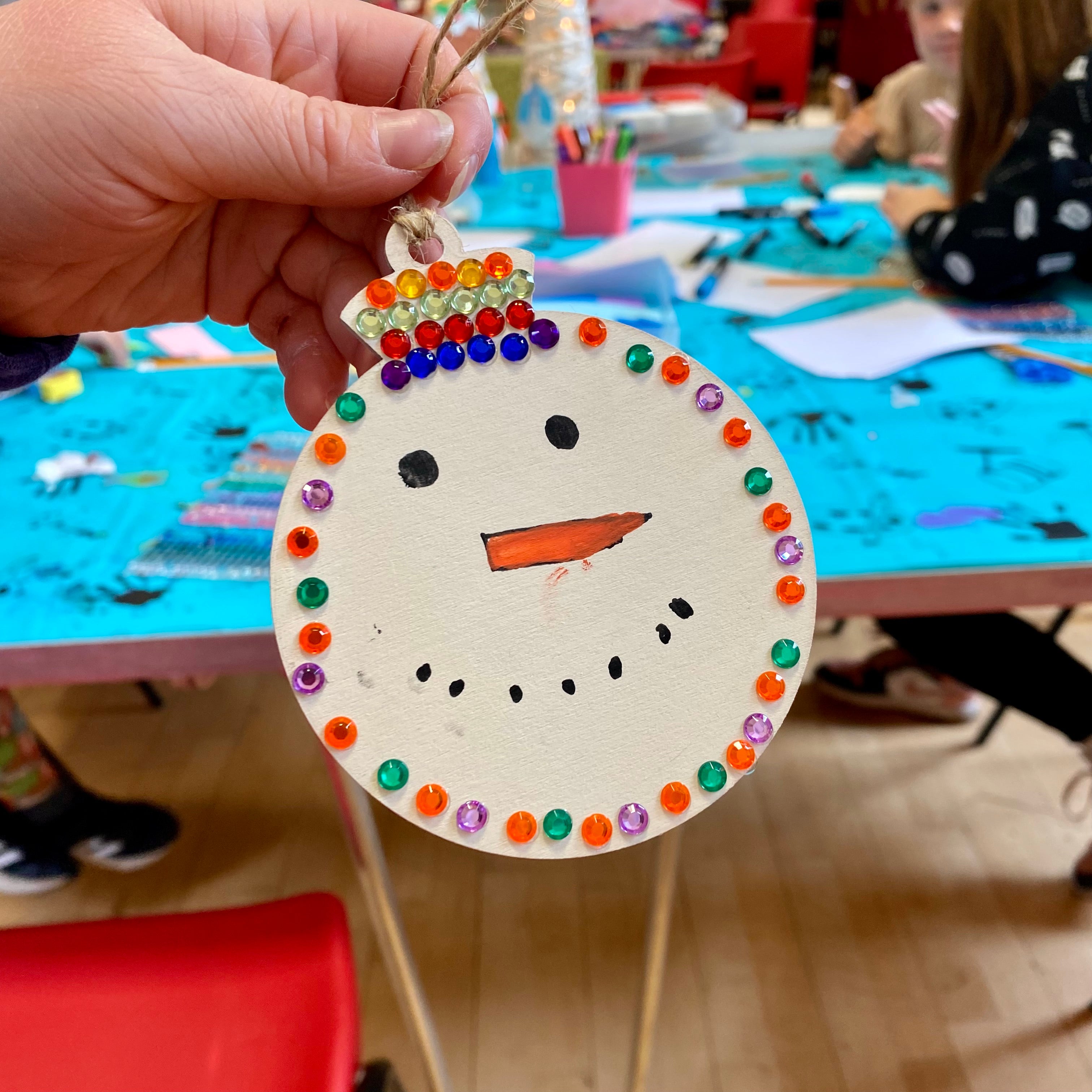 A snowman shaped crafted bauble with lots of sparkly gems on being held up. In the background is a workshop in full swing with other people sat around a table creating their own custom baubles.