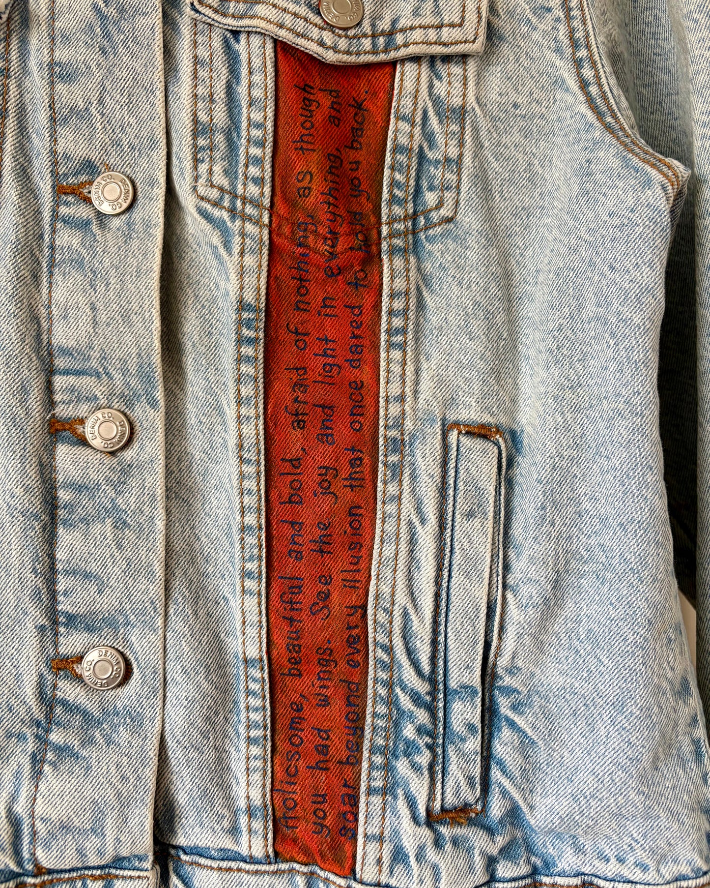 Close-up of hand-painted lettering on the front of a denim jacket, showing affirmations including ‘soar beyond every illusion that once dared to hold you back’ on an orange panel. Unique, upcycled wearable art.