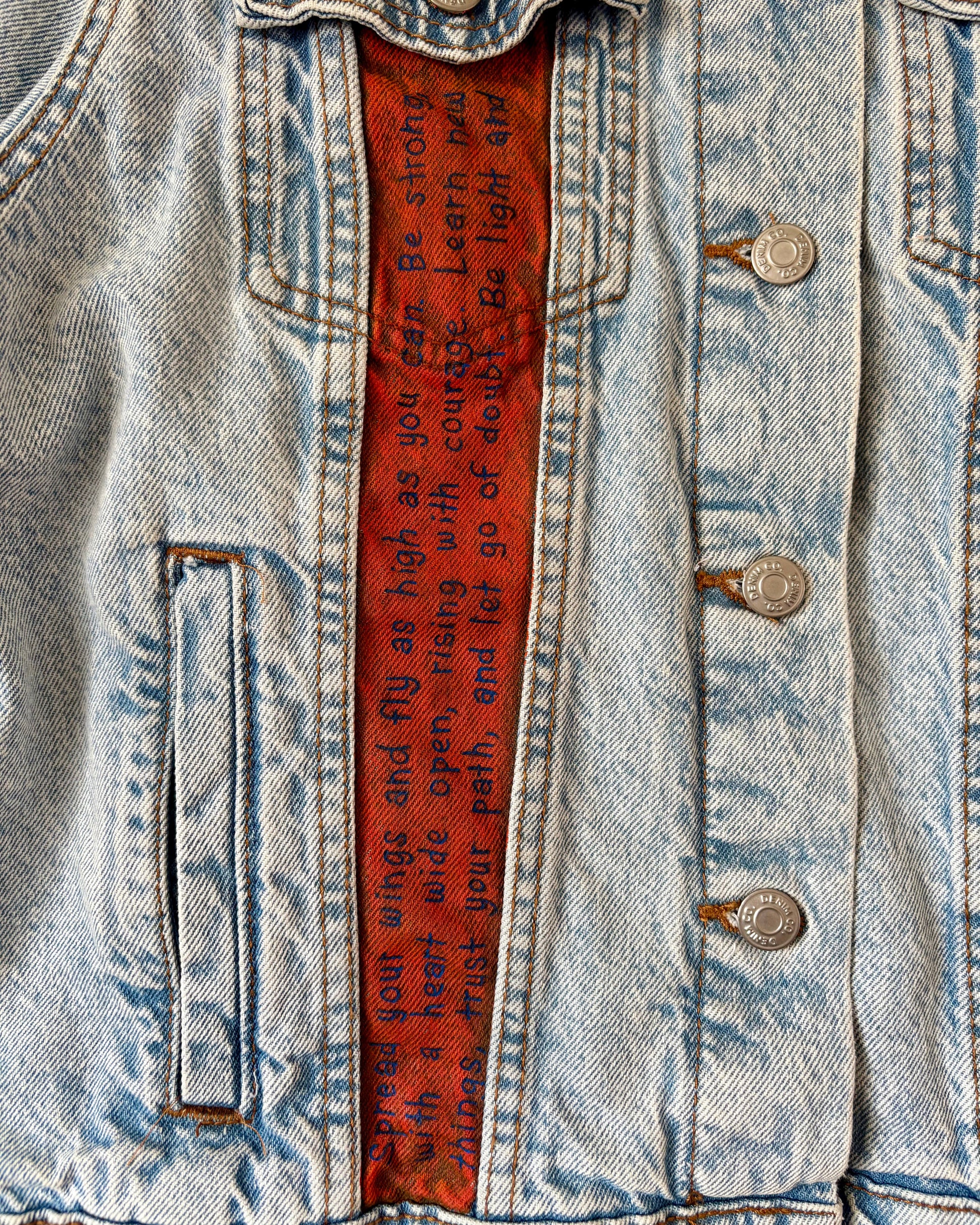 Close-up of hand-painted lettering on the front of a denim jacket, showing affirmations including ‘Spread your wings and fly as high as you can’ on an orange panel. Unique, upcycled wearable art.