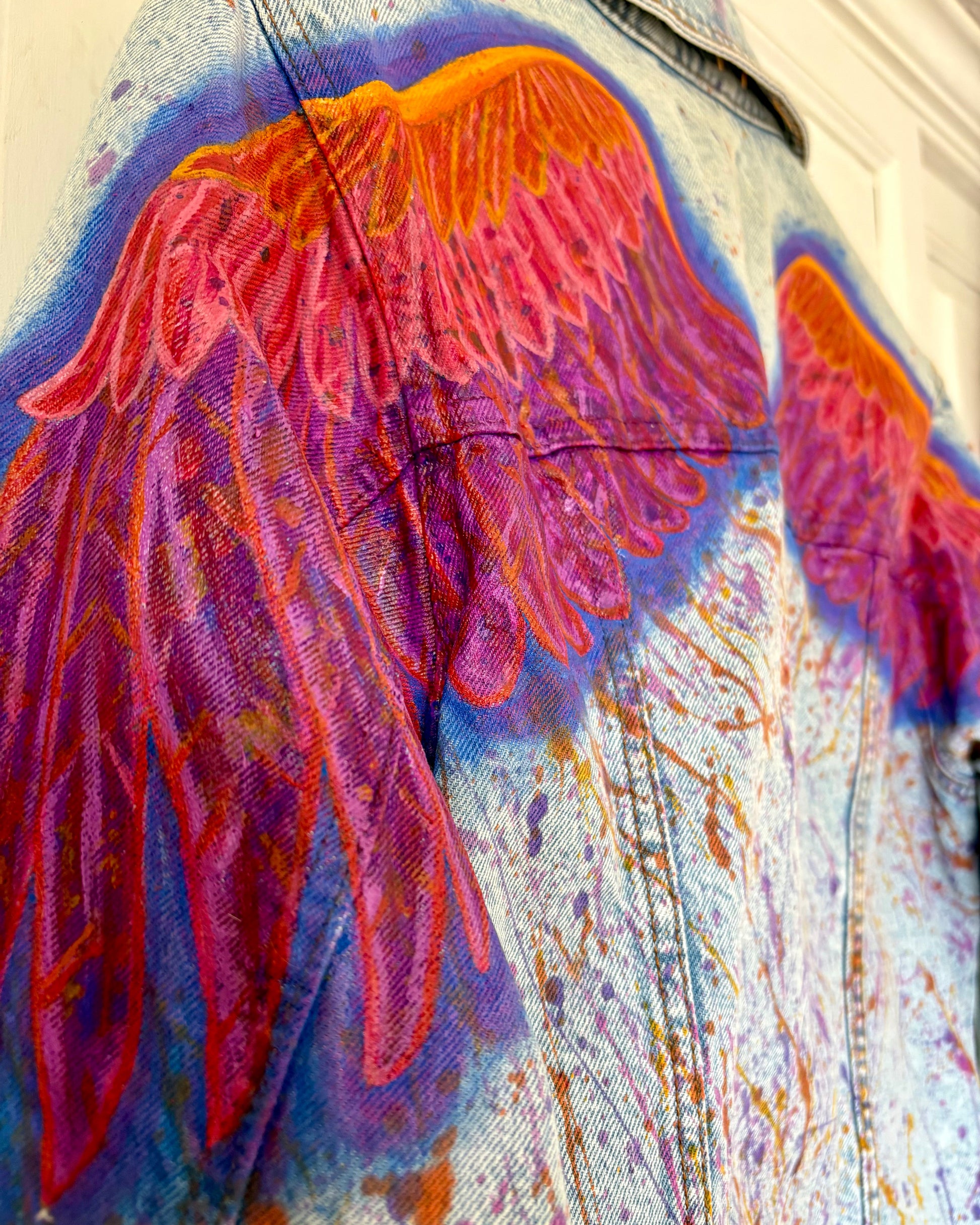 Close up of back of a hand-painted UK size 12 denim jacket featuring large bird wings in orange, pink, and purple painted across the shoulders and down the arms, with coordinating paint splashes extending under the wings and along the arms. Unique upcycled wearable art, one-of-a-kind design.