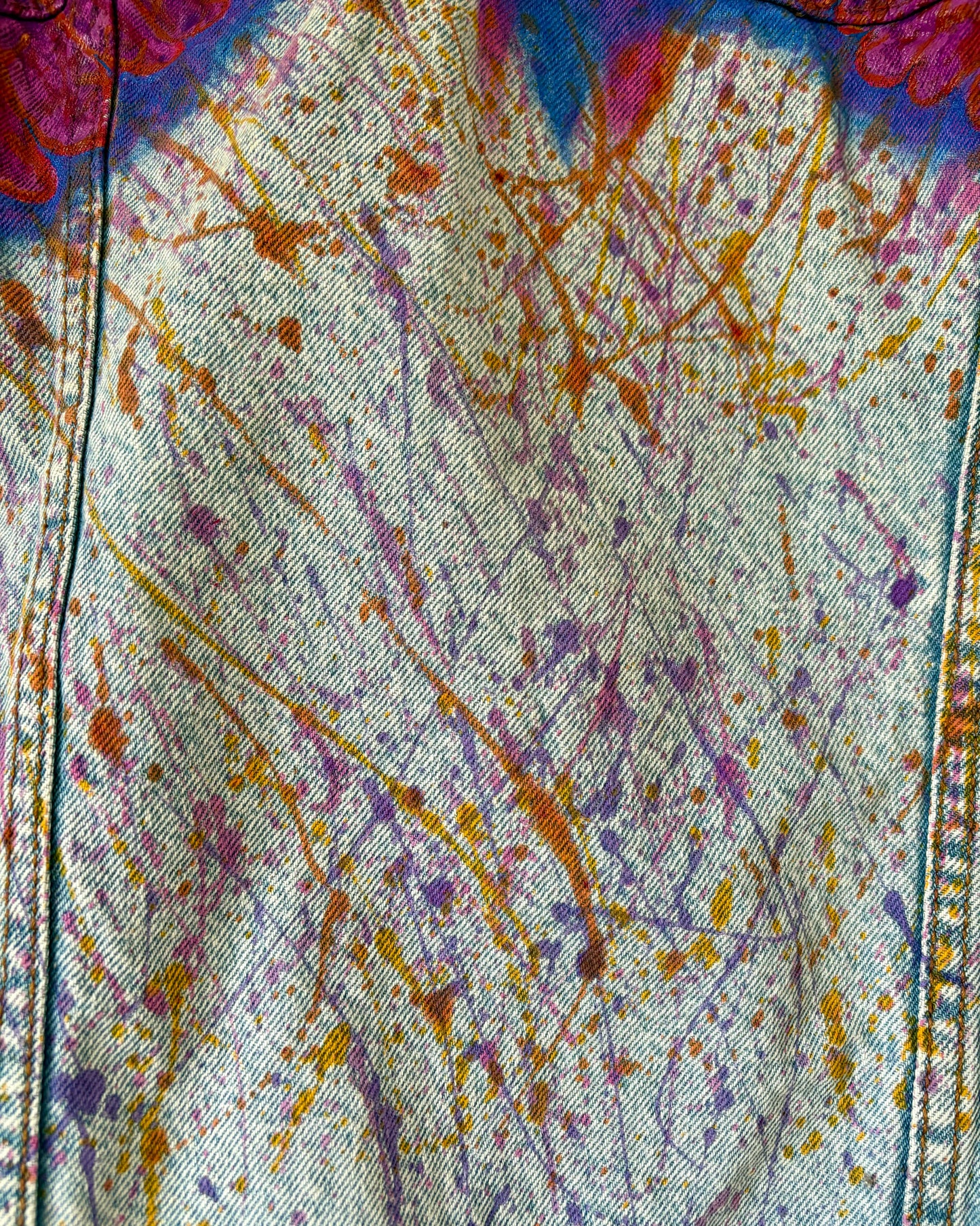 Close-up detail of hand-painted paint splashes on a denim jacket in orange, pink, and purple, flowing beneath the bird wings. Unique paint splat texture on one-of-a-kind upcycled wearable art.