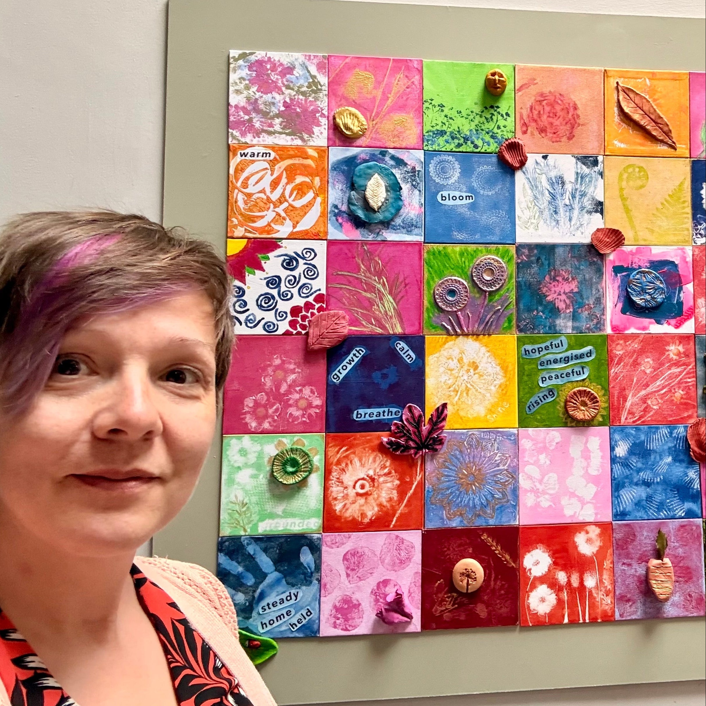 A woman with short pink-tinted hair stands beside a large framed colorful mosaic artwork made of many square canvas panels in bright hues like pink, blue, yellow, green, and orange, with clay nature inspired elements.