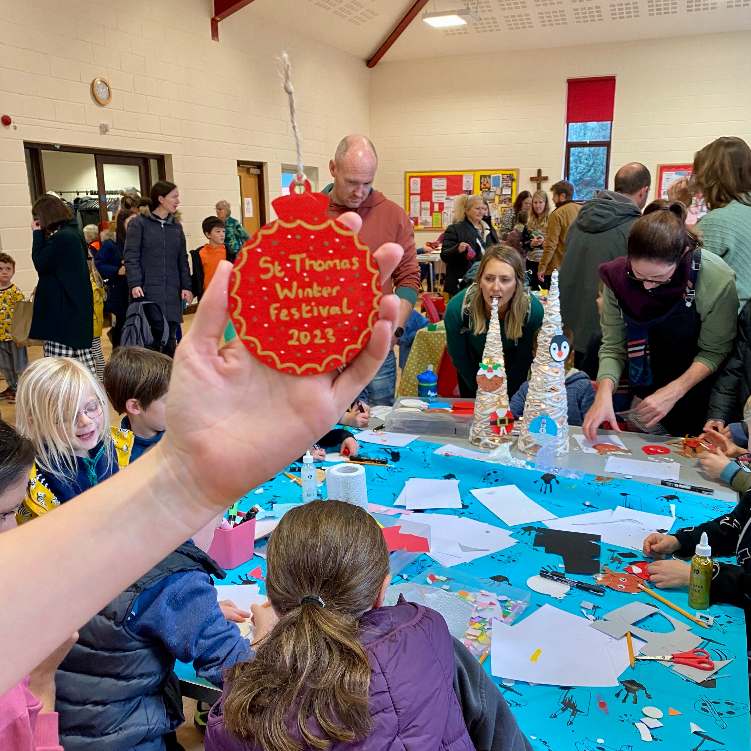 A red and gold painted bauble with the words 'St Thomas Winter Festival 2023' on held up in front of a crafting table where a workshop is in full swing with children sat around a table creating their own Christmas baubles
