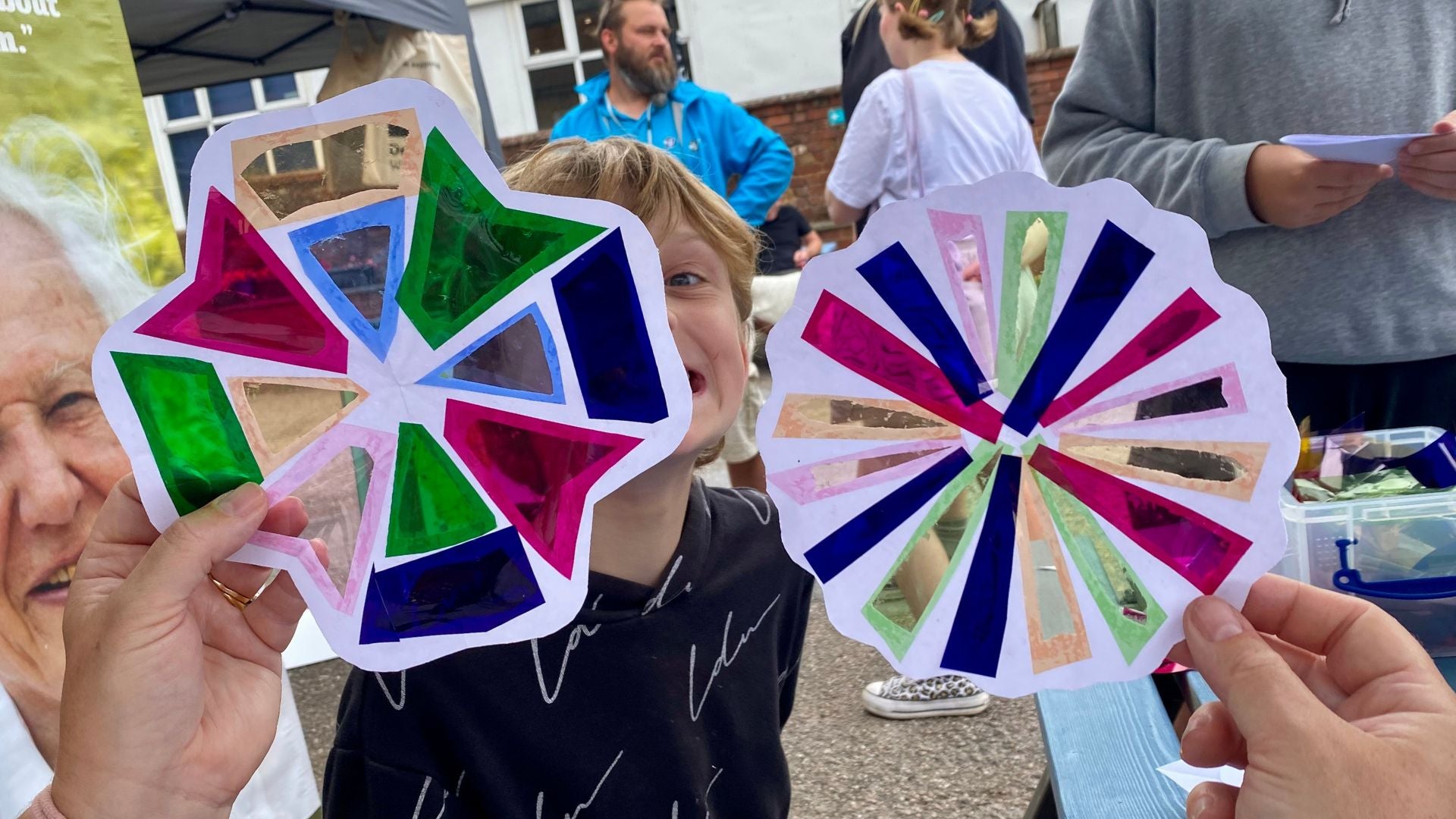 2 colourful flower shaped suncatchers being held up with a happy child behind. They are made from paper and coloured acetate. There are market stalls in the background.