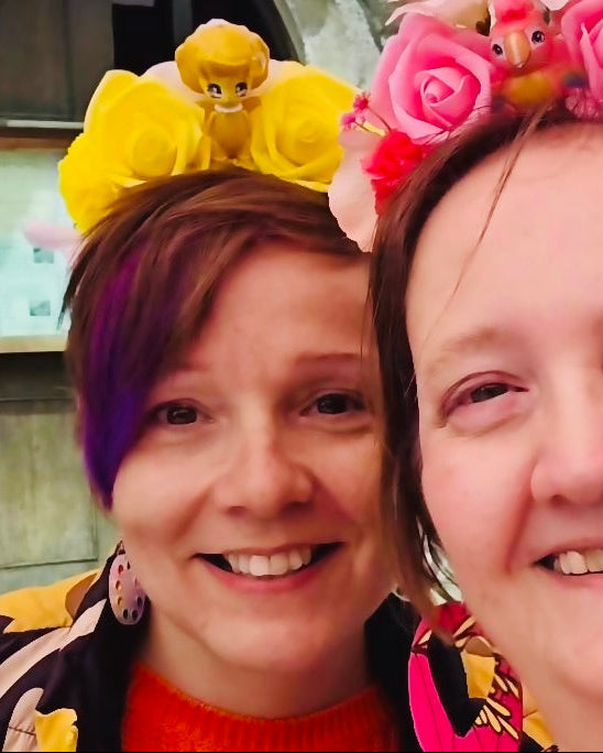 Two smiling women taking a selfie outdoors. The woman on the left wears a yellow flower crown with a cartoon character.