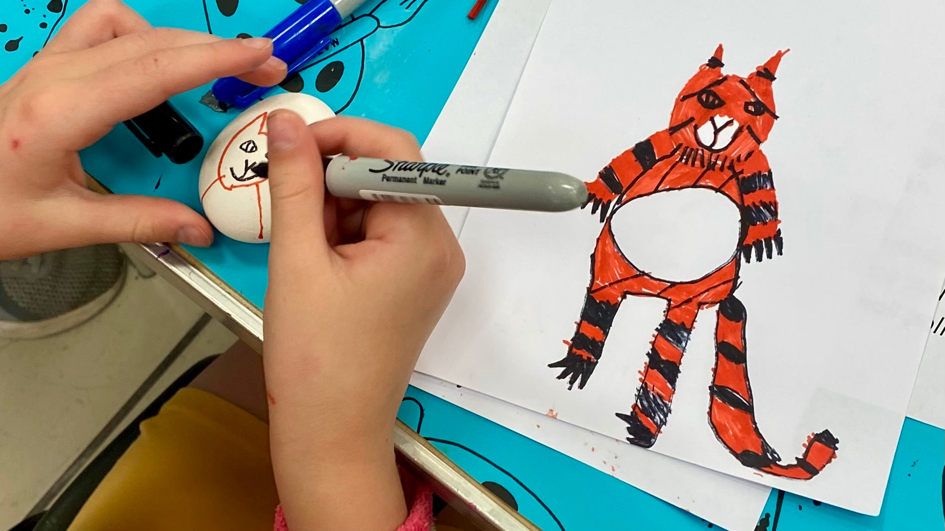 Child's hands drawing details of a tiger on a stone with a pen. Next to the stone is a drawing of a tiger which had been drawn by the child.