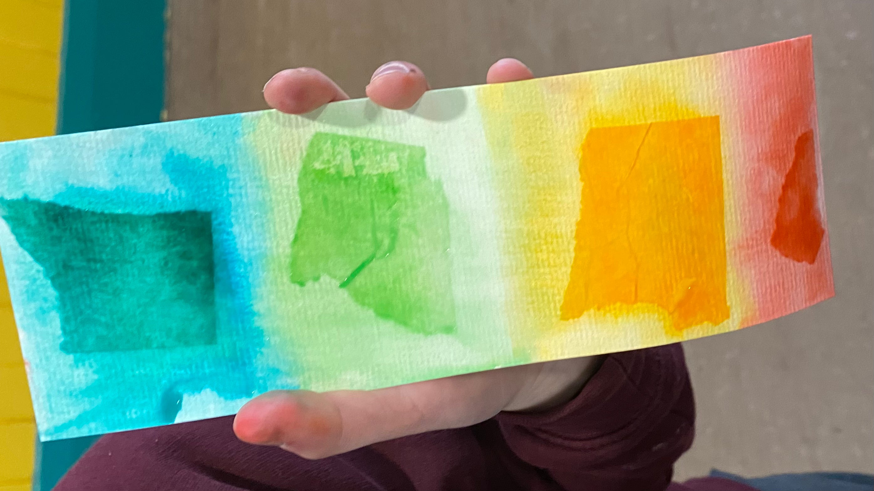 A hand holding a tissue paper printed textured card bookmark. Colours blue, green, yellow and red stripes blended into each other.