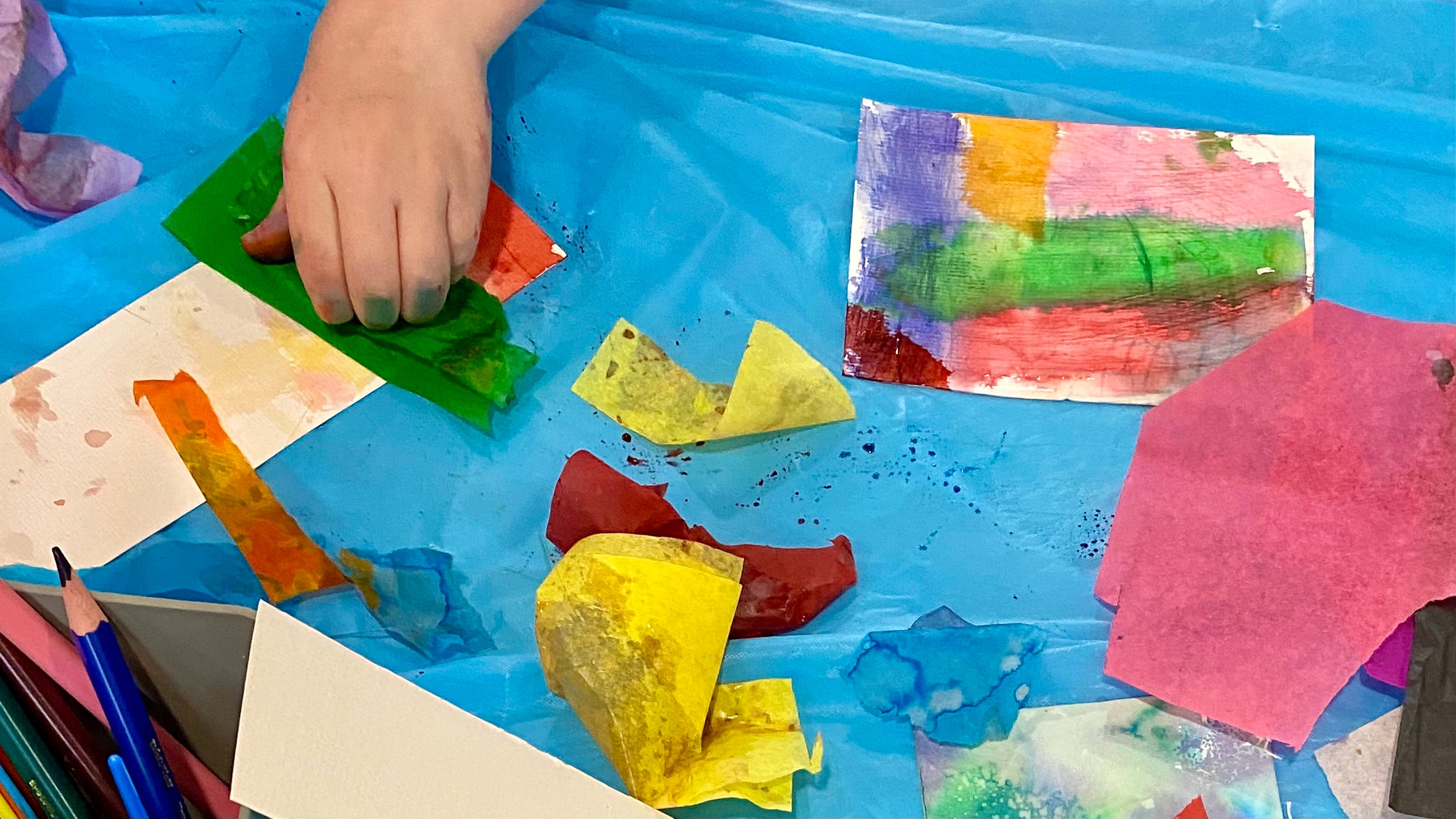 A top down view of a art table with a hand pressing green tissue paper onto a piece of wet card, surrounded by colourful tissue paper and art print creations