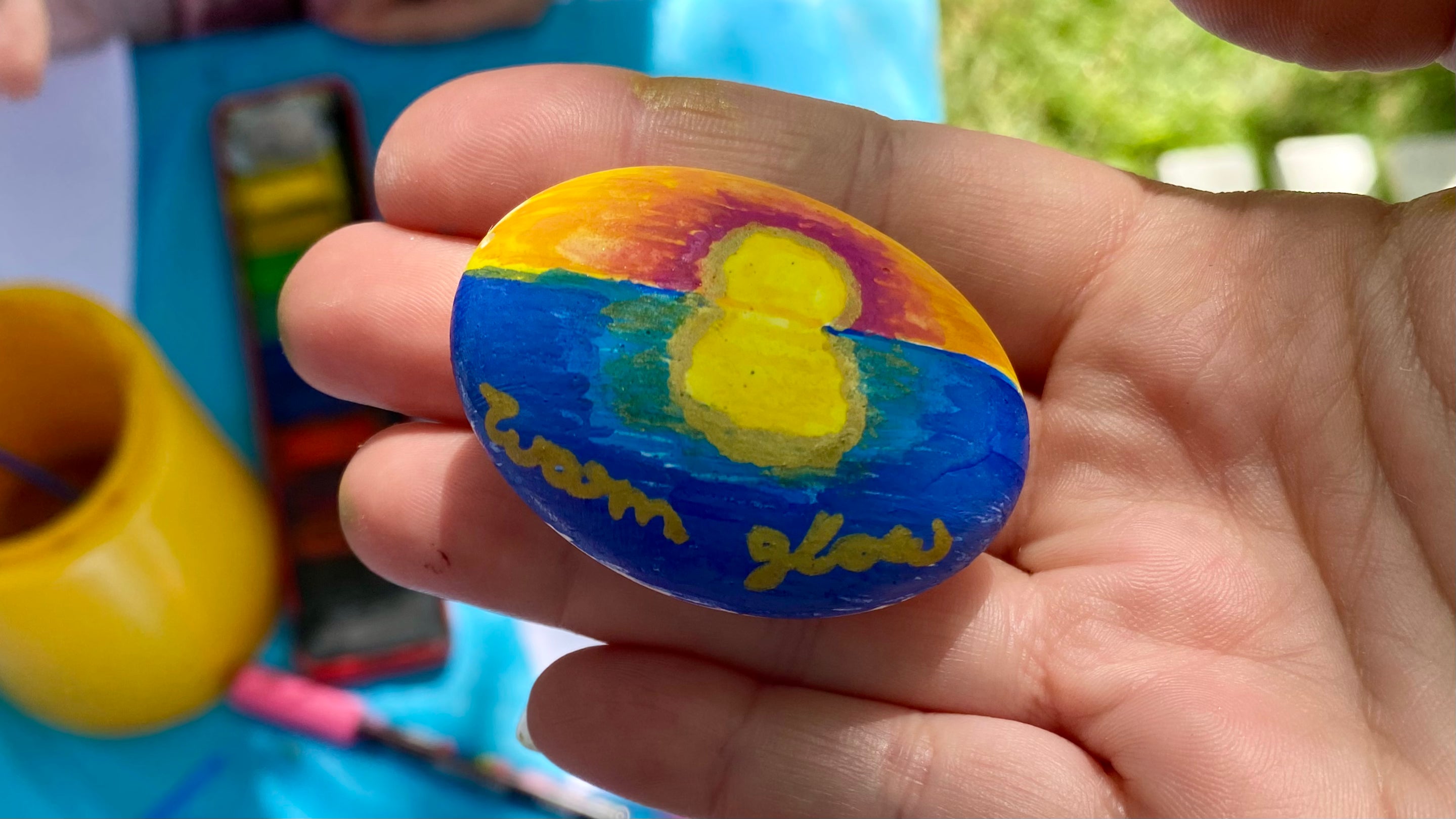 Small painted pebble held in a hand with a sunset painted on and the words Warm Glow written in gold