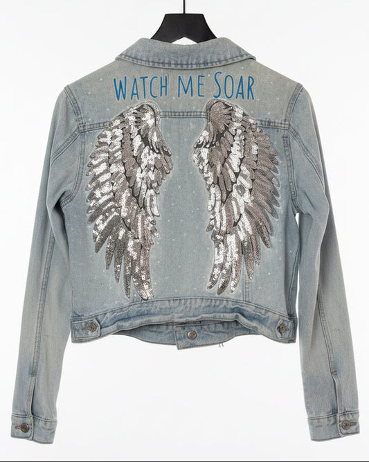 Denim jacket with sequin wings and 'Watch Me Soar' text painted on.