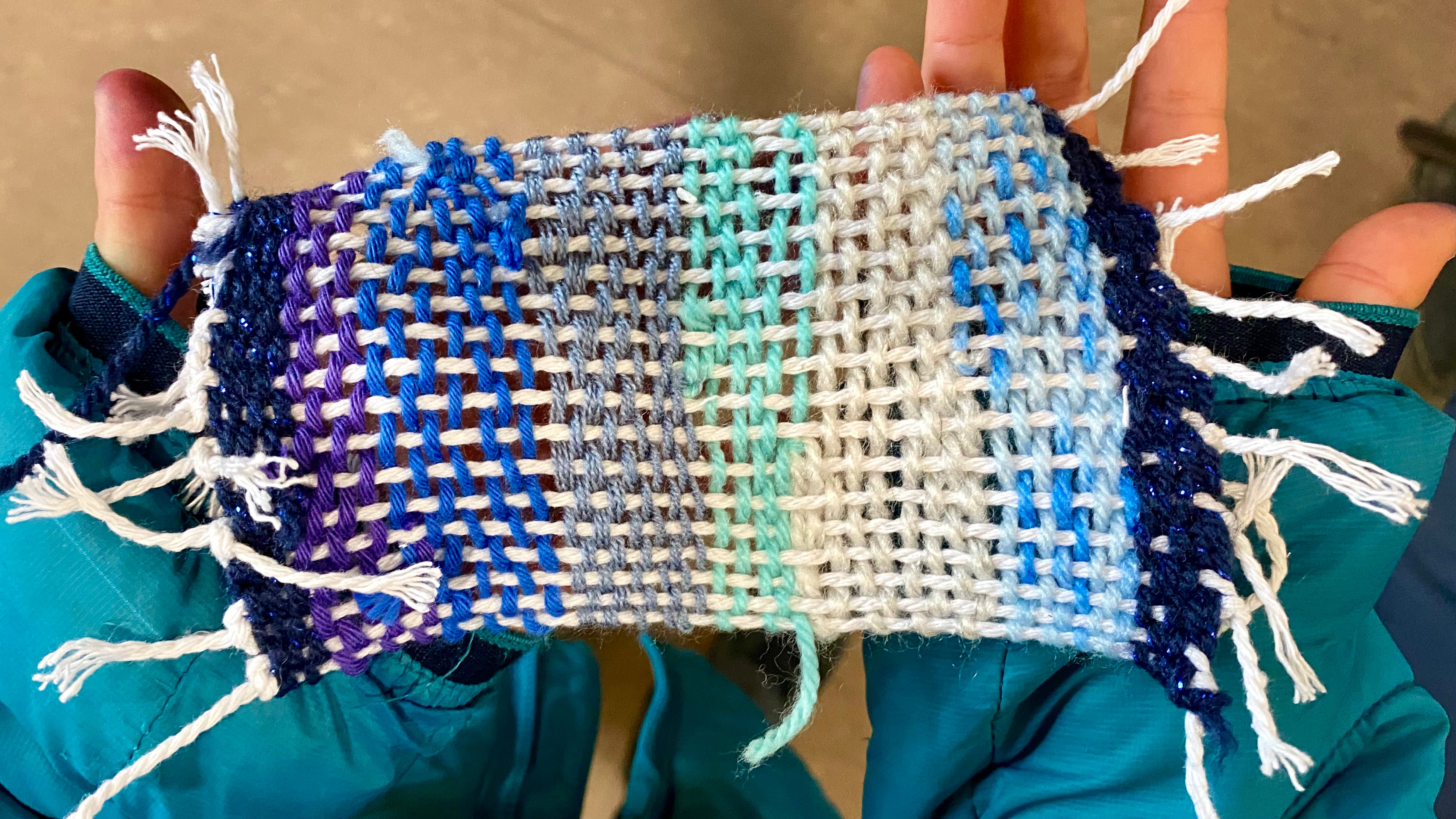 A pair of hands holding a finished weaving rectangle using scrap yarn. The colours are blues, teals, beige, grey and sparkly navy.
