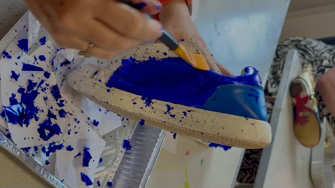 A person's hands applying metallic blue gold leaf onto a white leather shoe, surrounded by blue leaf flecks in a tray.