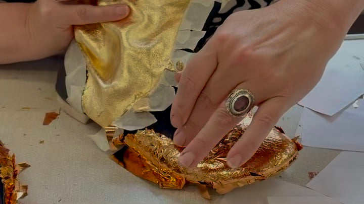 A close-up of a person's hands applying layers of shimmering gold and copper leaf onto a boot. 
