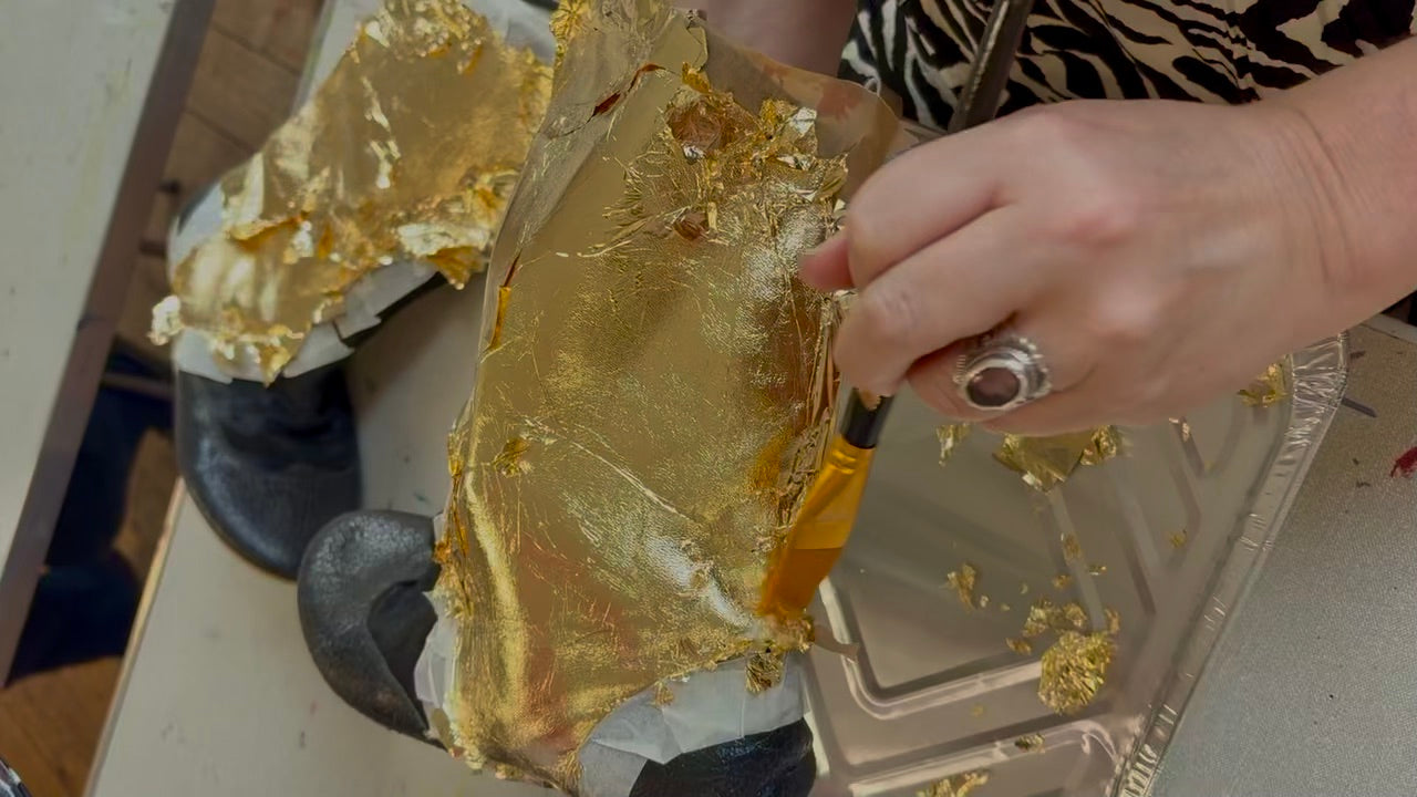 A close-up of a person's hand applying or smoothing metallic gold leaf onto a black shoe, with additional gold leaf and a tray of flakes visible on a tabletop.