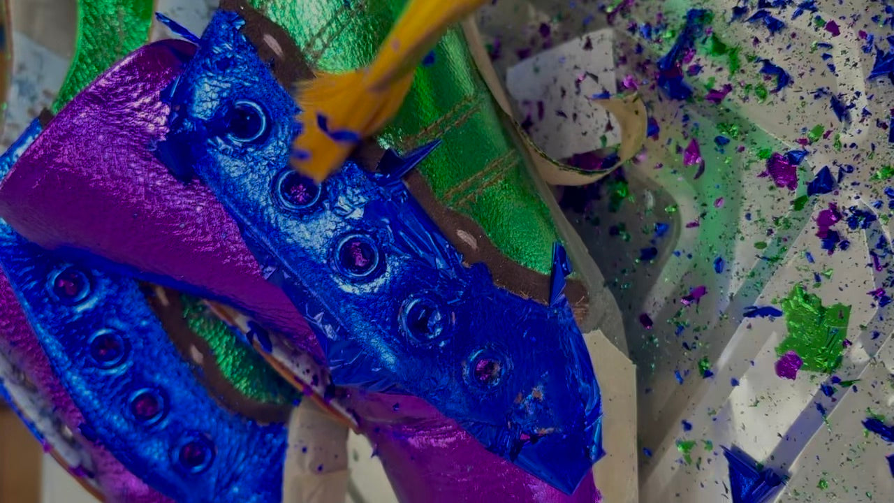 A close-up of a colorful, textured shoe covered in bright blue, green and purple metallic leaf, being smoothed out with a soft brush.