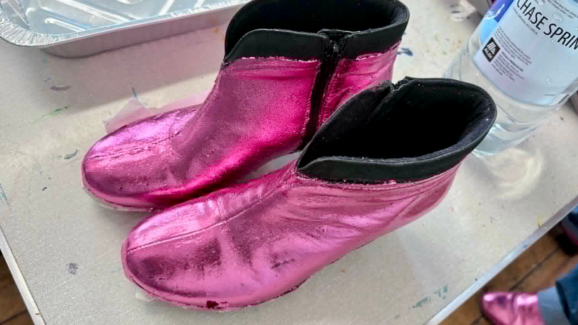 A pair of foiled metallic pink boots shown on a light-colored workbench.