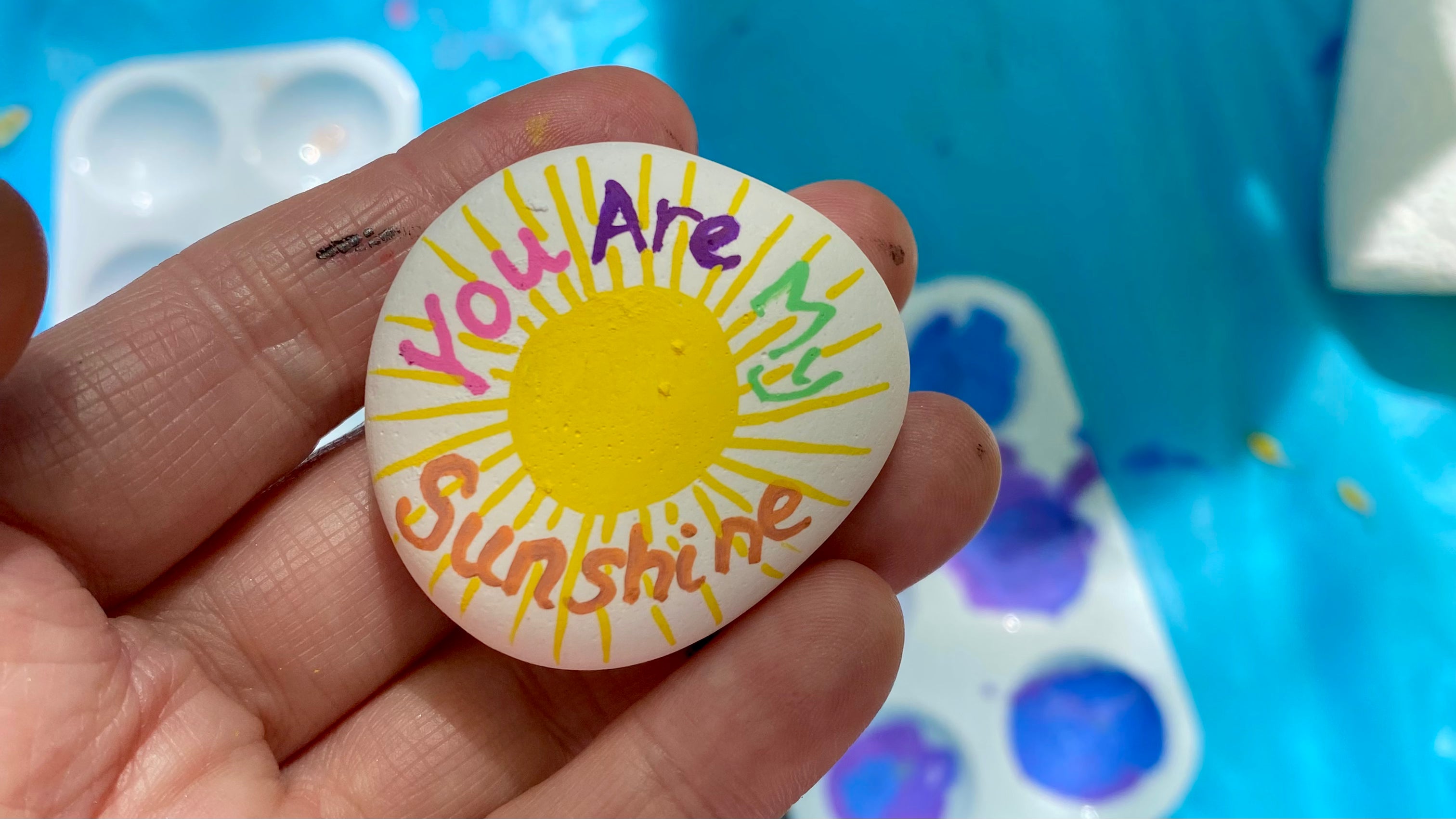 Hand holding a painted pebble with a yellow sunshine and the words You Are My Sunshine painted on in bright multicolours