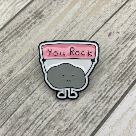 An enamel pin with a cartoonish design of a smiling rock wearing sunglasses and a sign that reads 'You Rock'. The pin is pink and grey with black outlines and a white background.
