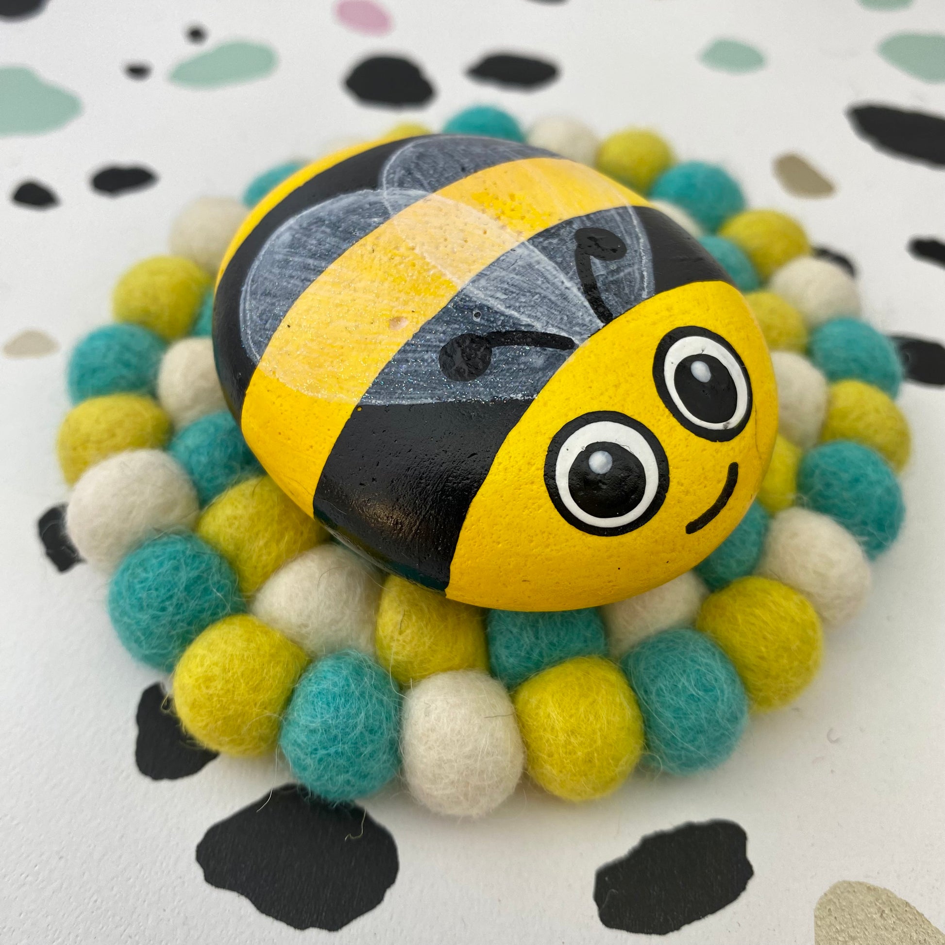 A Smooth Hand painted Yellow and Black Bee Pebble with lightly sparkling wings, and a cute face with big painted eyes. Sat on a felt mat or 'bug rug'.