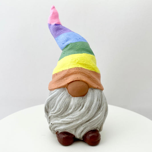 A small hand painted gonk statue with a pastel rainbow striped hat, light grey beard and brown shoes. 