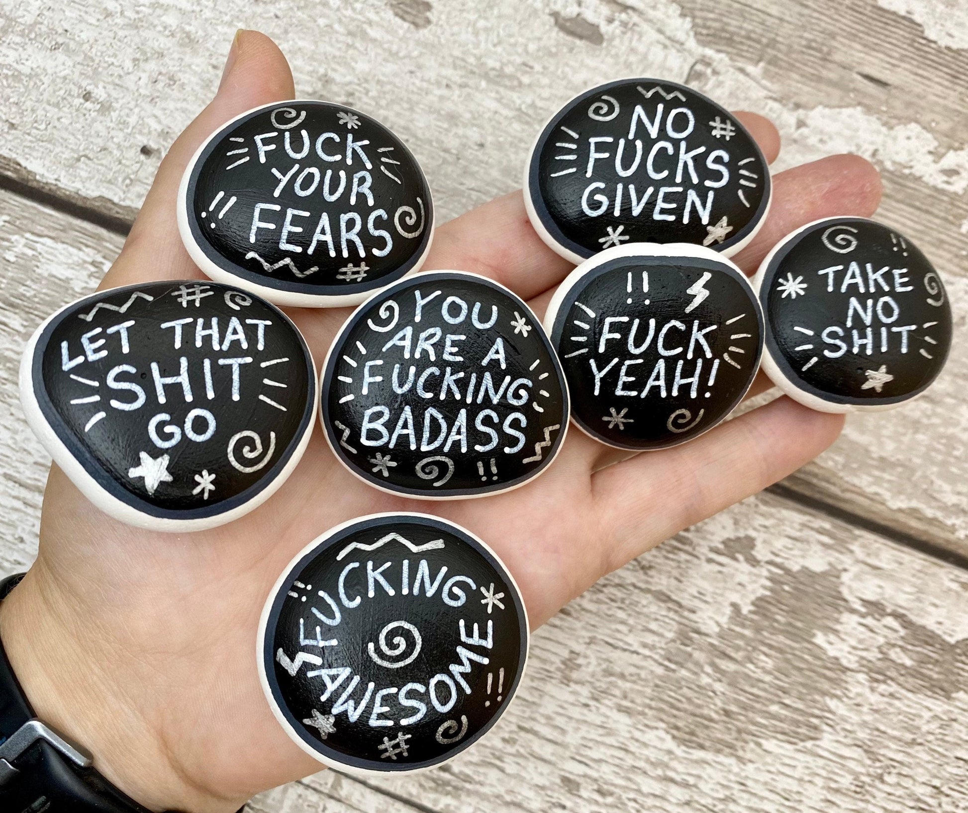 Hand painted Sweary Pebbles with rude affirmation messages. Silver words on a black background