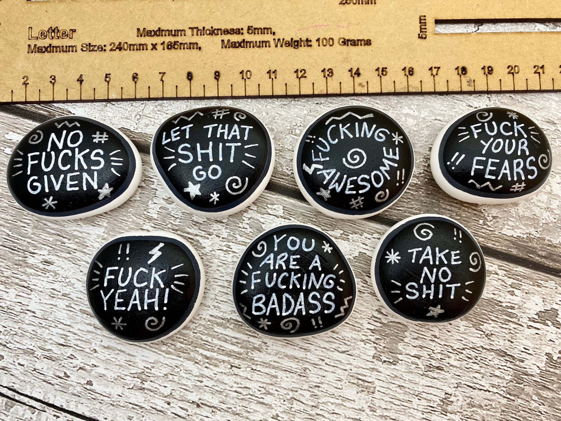 Hand painted Sweary Pebbles with rude affirmation messages next to a ruler. Silver words on a black background.