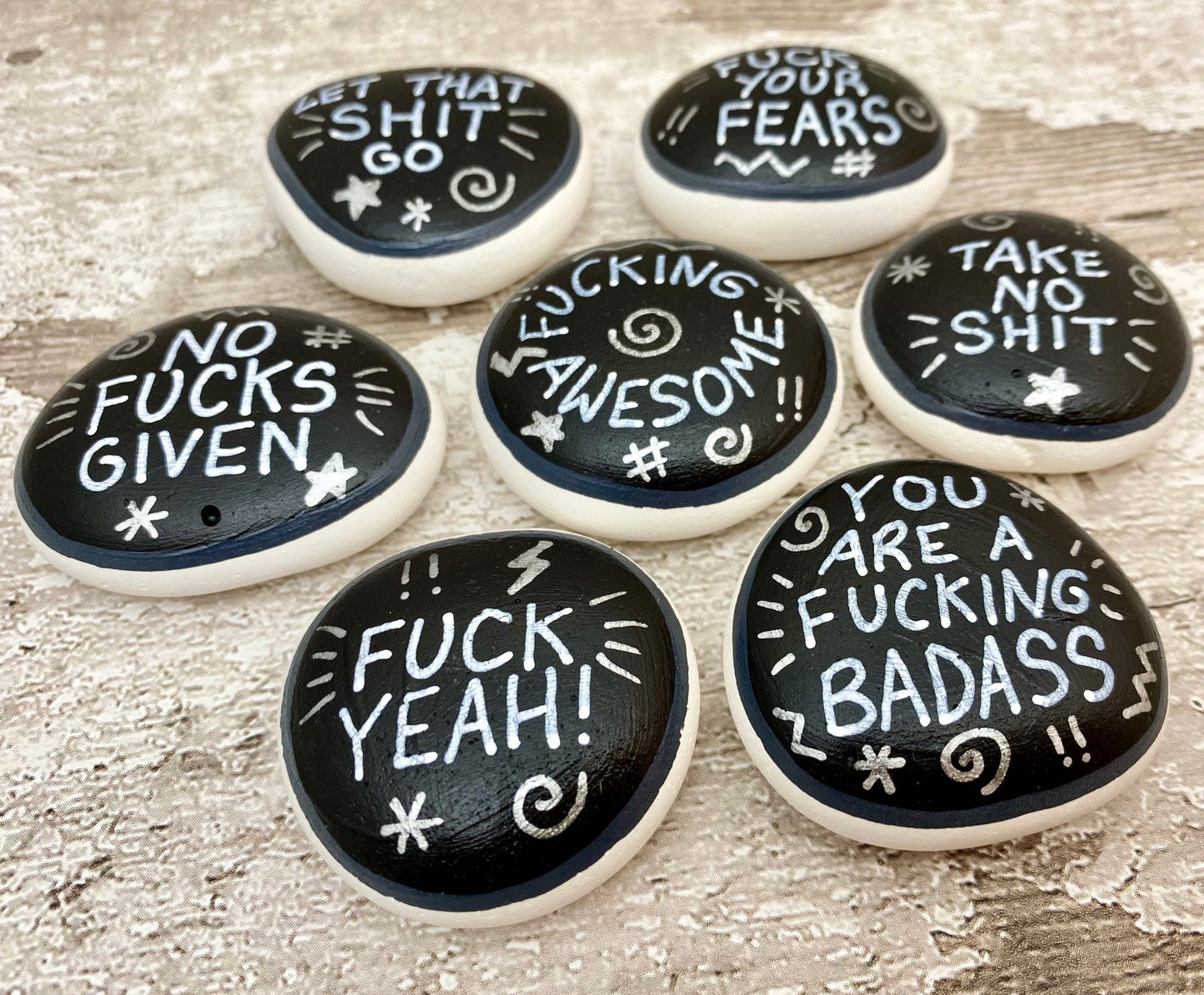 Hand painted Sweary Pebbles with rude affirmation messages. Silver words on a black background